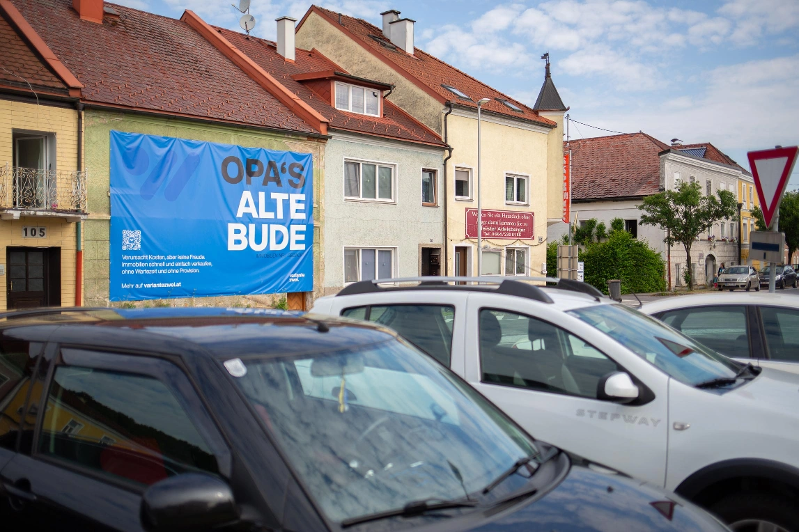 A street scene with parked
                cars and a large blue banner on a building advertising 'opa's
                alte bude' with german text.